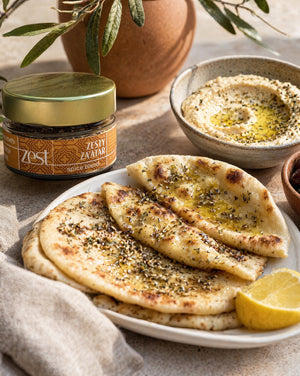 A jar of Zesty Za'atar spice blend is shown with a plate of za'atar-dusted flatbread, a bowl of hummus drizzled with olive oil, and a lemon wedge on a light-colored surface.